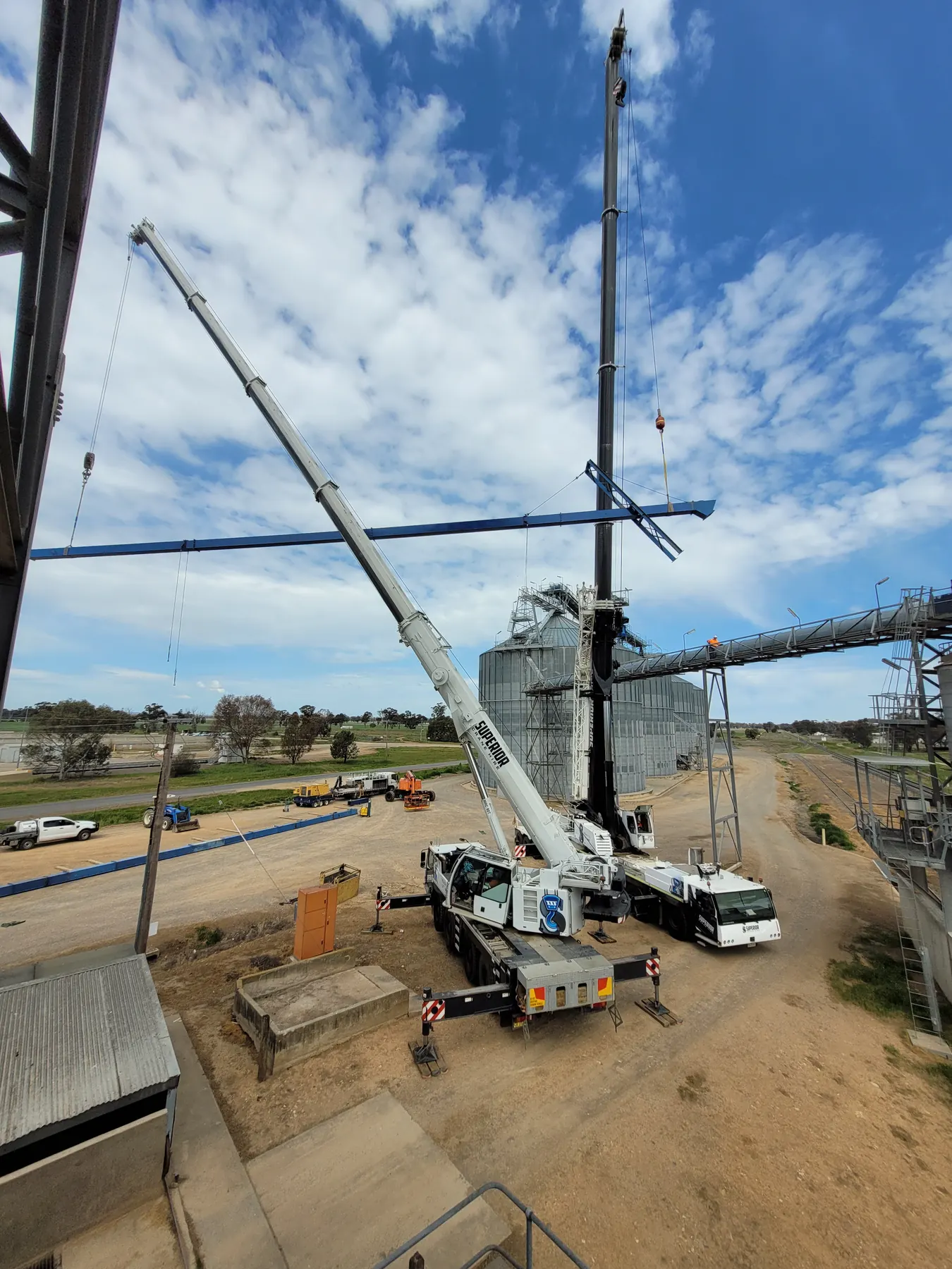 Two cranes lifting a long steel beam at a grain facility